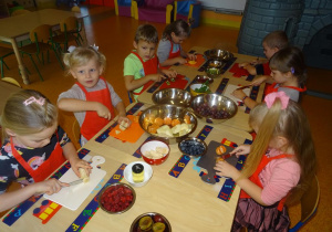 Grupa dzieci siedzi przy stole, w ręku trzymają noże, którymi kroją owoce na desce.